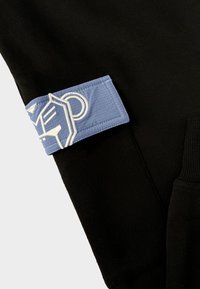 Zwarte sweatpants hebben een lichtblauwe patch met witte geborduurde grafische details. De stof lijkt zacht met een gladde textuur.