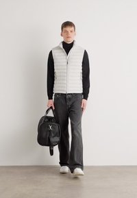 Vit vadderad väst över en svart polotröja, gråjeans, vita sneakers och bärande en svart väska. Enkelt design med en mjuk textur.