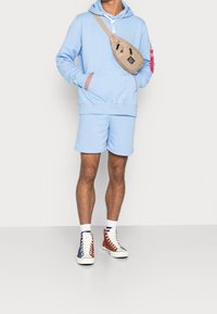 Felpa blu chiaro con tasca anteriore, pantaloni corti abbinati, borsa a tracolla beige e sneakers alte multicolori con trame contrastanti.