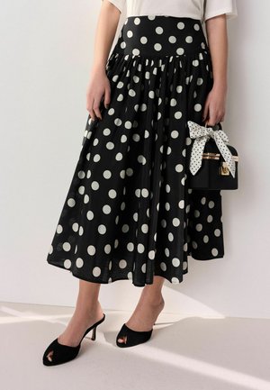Frau trägt einen schwarzen Midirock mit großen weißen Polka-Dots, schwarze offene Schuhe mit Absatz, hält eine schwarze Handtasche mit weißer gepunkteter Schleife.