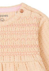 Perzikkleurige jurk met een smock-textuur en roze stikselaccenten. Heeft een ronde halslijn en een bloemenknop op de schouder.