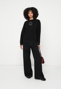 Černá mikina s kulatým výstřihem, zdobená úsměvem uprostřed a kombinovaná s černými širokými kalhotami.