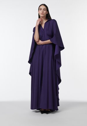 Mujer de pie con un vestido largo color púrpura intenso con mangas tipo capa y zapatos negros puntiagudos sobre un fondo blanco liso.
