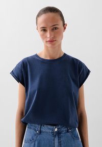 T-shirt in cotone blu navy con maniche corte arrotolate e scollatura a girocollo, con un piccolo logo ricamato sul petto. Indossato con jeans di denim.
