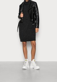 Svart läderbikerjacka över en mörkgrå stickad klänning, kombinerad med vita kraftiga snörstövlar. Slät struktur, figurnära design och lager-på-lager stil.