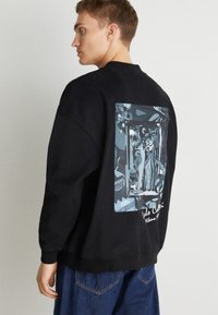 Zwarte sweatshirt met een grote grafische print op de rug met blauwe en witte illustraties. De stof lijkt zacht en textuur te hebben.