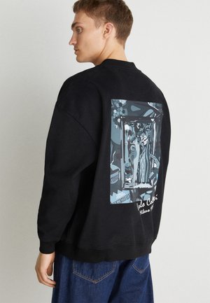 Sweatshirt noir avec un grand imprimé graphique dans le dos comportant des illustrations bleues et blanches. Le tissu semble doux et texturé.