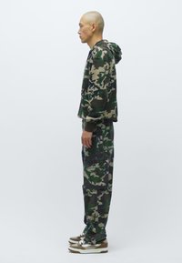 Vīrietis stāv profilā, tērpies zaļā un brūnā kamuflāžas kapucē, atbilstošās cargo biksēs un brūnos un baltos sporta apavos.