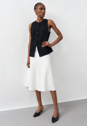 Žena stojaca s rukou na boku, oblečená v čiernom bezrukávovom zapínanom topu, bielej midi sukni a čiernych lodičkách s špicatou špičkou na jednoduchom pozadí.