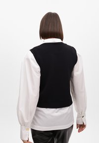 Gilet nero lavorato a maglia sopra una camicia bianca con bottoni e maniche a sbuffo. L'outfit presenta un design semplice e dettagli con bottoni sulle spalle.