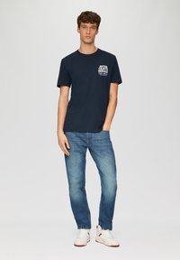 Marineblauw katoen t-shirt met een witte grafische print op de borst, gecombineerd met vervaagde blauwe spijkerbroeken en witte sneakers.