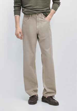 Uomo che indossa pantaloni beige a gamba dritta con un maglione verde, con la mano destra in tasca e scarpe di pelle marrone.