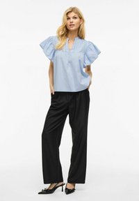 Gaiši zila īsām piedurknēm blūze ar volāniem, V-veida kakla izgriezumu un vertikālām šuvēm, apvienota ar melnajiem platiem biksēm un papēžiem.