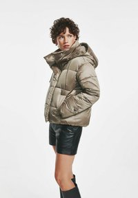 Padded, metaliczna oliwkowa kurtka z kapturem, z zamkiem błyskawicznym, bocznymi kieszeniami i elastycznymi mankietami. Nosić na czarnych skórzanych szortach.
