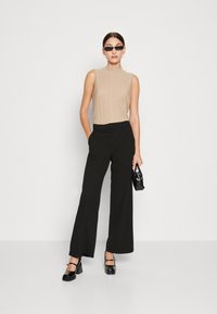 Haut sans manches en maille beige, col montant. Pantalon large noir, matériau lisse, accents à deux boutons. Sac à main noir et chaussures à talons.
