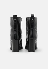 Botas de cuero negras a los tobillos con un acabado suave, tacones rectangulares y cremalleras en los lados exteriores. El detalle trasero incluye pestañas con bucles.