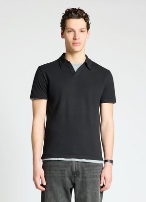 Young man with curly hair wearing a black short-sleeve polo shirt over a light gray undershirt and dark gray jeans.