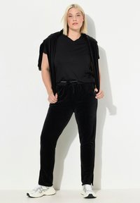 Zwarte fluwelen joggers en een bijpassend los V-hals t-shirt, gelaagd met een korte zwarte open voorkardigan. Witte sportschoenen maken de outfit compleet.