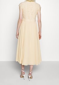 Kvinna iklädd en beige spetsklänning med plisserad kjol och vita högklackade sandaler, stående mot en neutral vit bakgrund.