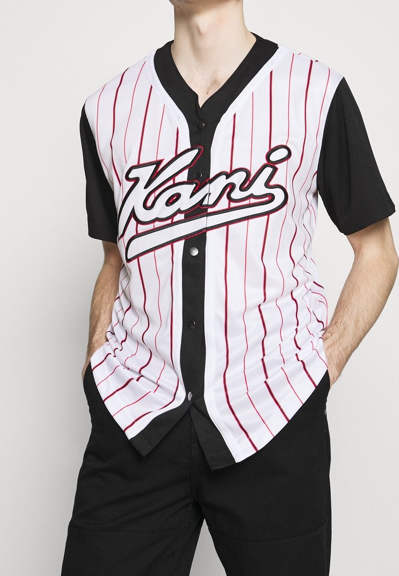 Maillot de baseball blanc avec des rayures verticales rouges et des manches courtes noires. Présente une fermeture à boutons sur le devant et un grand logo "Kani" sur la poitrine.