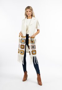 Häkel-Schal in Creme mit mehrfarbigen Sechseckmustern und fransigen Kanten. Das Model trägt einen hellen Pullover und dunkle Jeans, mit braunen Stiefeletten.
