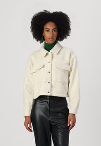 Cropped, cream-colored fluffy jacket with snap buttons, two chest pockets, and a collared neck. Paired with black leather pants.