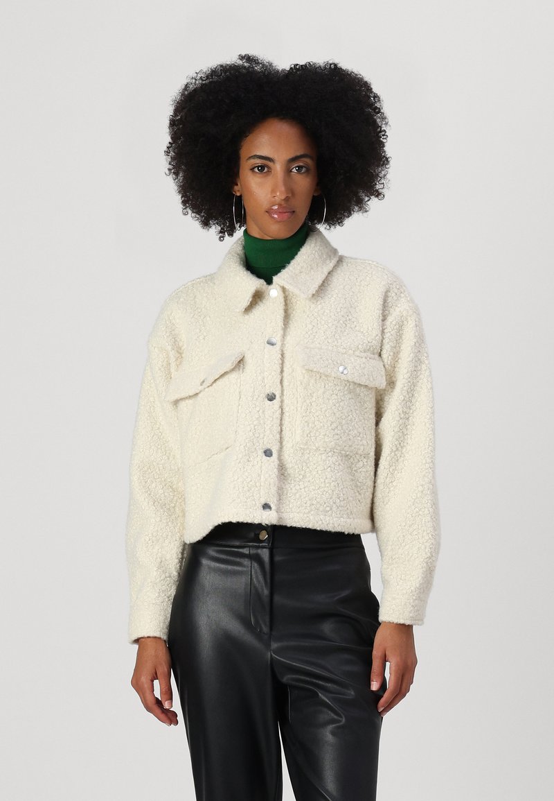 Cropped, cream-colored fluffy jacket with snap buttons, two chest pockets, and a collared neck. Paired with black leather pants.