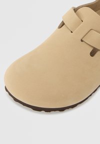 Beige suede slip-on kenkiä, joissa on pyöreä kärki, ja jotka sisältävät yhden hihnan ylhäällä sekä tekstuurituloksen ruskea pohja.