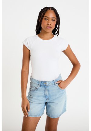 Jeune femme aux cheveux tressés portant un haut blanc ajusté à manches courtes et un short en denim bleu clair, debout avec une main dans la poche.