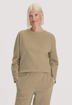 Frau mit glatten blonden Haaren trägt beige geripptes Langarmshirt und passende Hose, steht mit den Händen in den Taschen vor einfachem Hintergrund.