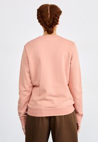 Rosa bomullssweater med lange ermer, rund hals og ribbede mansjetter, sammen med brune bukser. Baksiden har en jevn tekstur.