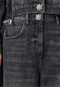 Zwarte denimjacket met een cropped ontwerp, voorzien van twee decoratieve bloemenknopen, één zak en rafelige randen bij de zoom.