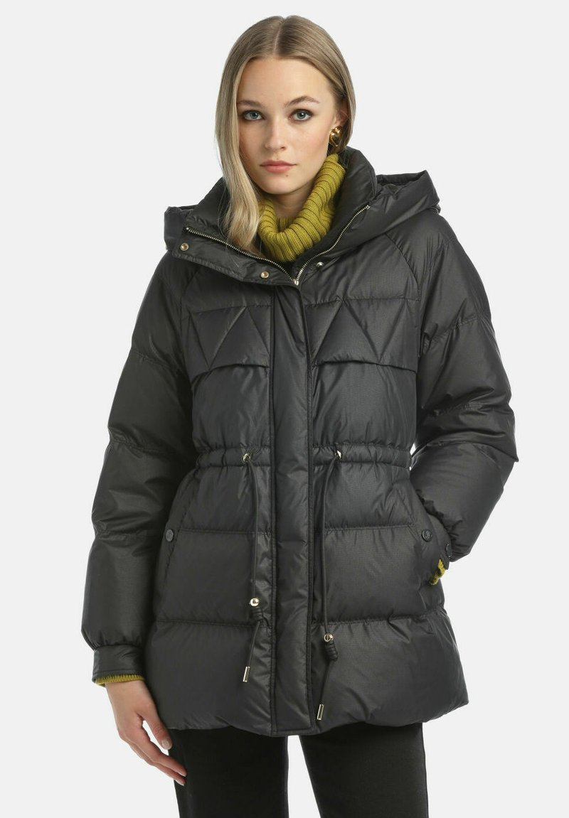 Črna izolirana puhovka s kapuco, z diamantskim prošivom, zategovalnim pasom in zlatimi dodatki. Oblečena čez zeleno pleteno jopico.