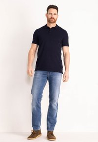 Námořnické polo tričko s krátkými rukávy, žebrovaným límečkem a tříknoflíkovým zapínáním; nošeno s světlými modrými džíny a hnědými semišovými botami.