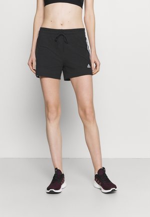 Personne portant un short Adidas noir avec des bandes blanches et des chaussures de sport foncées, debout devant un fond clair uni.