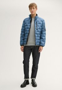 Jasnoniebieska kurtka puchowa z pikowanego materiału, z wysokim kołnierzem i kieszeniami na zamek, noszona na szarym swetrze i ciemnych dżinsach.