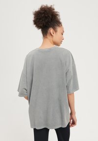 Kvinna med lockigt hår i en hög puff, klädd i en lös grå kortärmad tröja och svarta byxor, sedd från sidan med huvudet vänt åt höger.