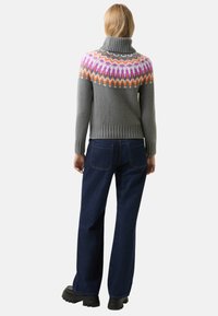 Osoba s blond vlasy, oblékající šedý rolák s růžovým, oranžovým a bílým vzorem, tmavě modré džíny a černé masivní boty, otočená zády.