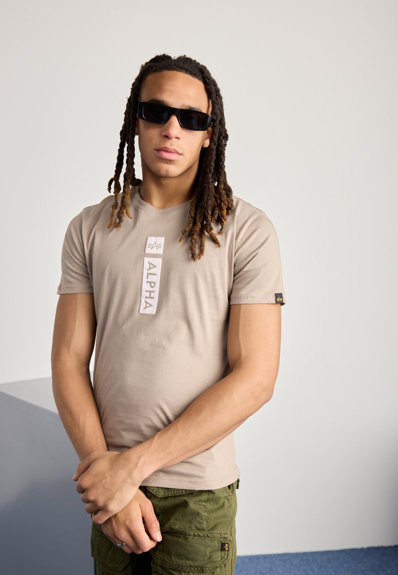 Beige t-shirt med kortermer, med "ALPHA" logo, rund halsudskæring og et lille mærke på ærmet. Bærer cargo-bukser og solbriller.