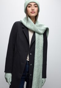 Svart ullkappa, ljus beige topp, denimjeans, grön stickad halsduk, matchande mössa och handskar, alla med en strukturerad, ribbad design.