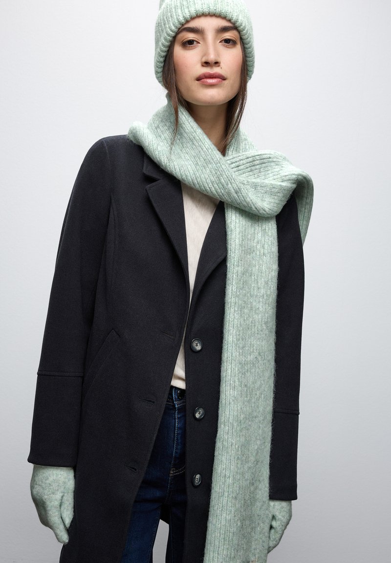 Svart ullkappa, ljus beige topp, denimjeans, grön stickad halsduk, matchande mössa och handskar, alla med en strukturerad, ribbad design.