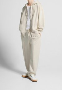 Ljusbeige huhoodtröja med framdragkedja, tillsammans med matchande sweatpants. Båda plaggen är gjorda av ett slätt, mjukt material.