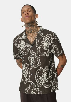 Homme avec des tatouages au cou et au bras, portant une chemise noire à manches courtes avec de grands motifs floraux blancs, debout les mains dans le dos.