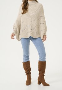 Béžový oversized pletený pončo s texturovaným vlnitým vzorem, spojený se světle modrými skinny džíny a hnědými kozačkami až po kolena. Viditelné detaily texturované příze.
