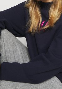 Laivastonsininen collegepaita, jossa on vaaleanpunainen kirjailu, pitkät hihat ja rento istuvuus. Yhdistetty vaaleanharmaisiin teksturoituihin collegehousuihin.