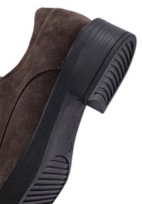 Chaussure en daim brun avec un bout arrondi et une semelle en caoutchouc noir texturé présentant des rainures horizontales pour l'adhérence et le style.