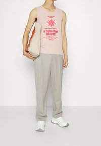 Person wearing a pink sleeveless top with red text, beige trousers, white sneakers, holding a beige and orange clutch bag against a white background.