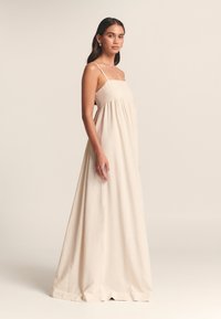Maxi suknelė šviesiai smėlio spalvos, su prigludusiu liemeniu, plonomis petnešėlėmis ir laisvai krentančia sijono dalimi su raukiniais tekstūrai.