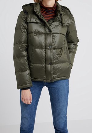 Osoba ubrana w zieloną, pikowaną kurtkę puchową na brązowo-czarnym pasiastym golfie i niebieskich jeansach, stojąca z jedną ręką w kieszeni.