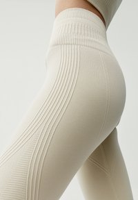 Béžové legíny s vysokým pásom vyrobené z textúrovaného pleteného materiálu s diagonálnymi a horizontálnymi rebrovanými vzormi po stranách nohavíc.